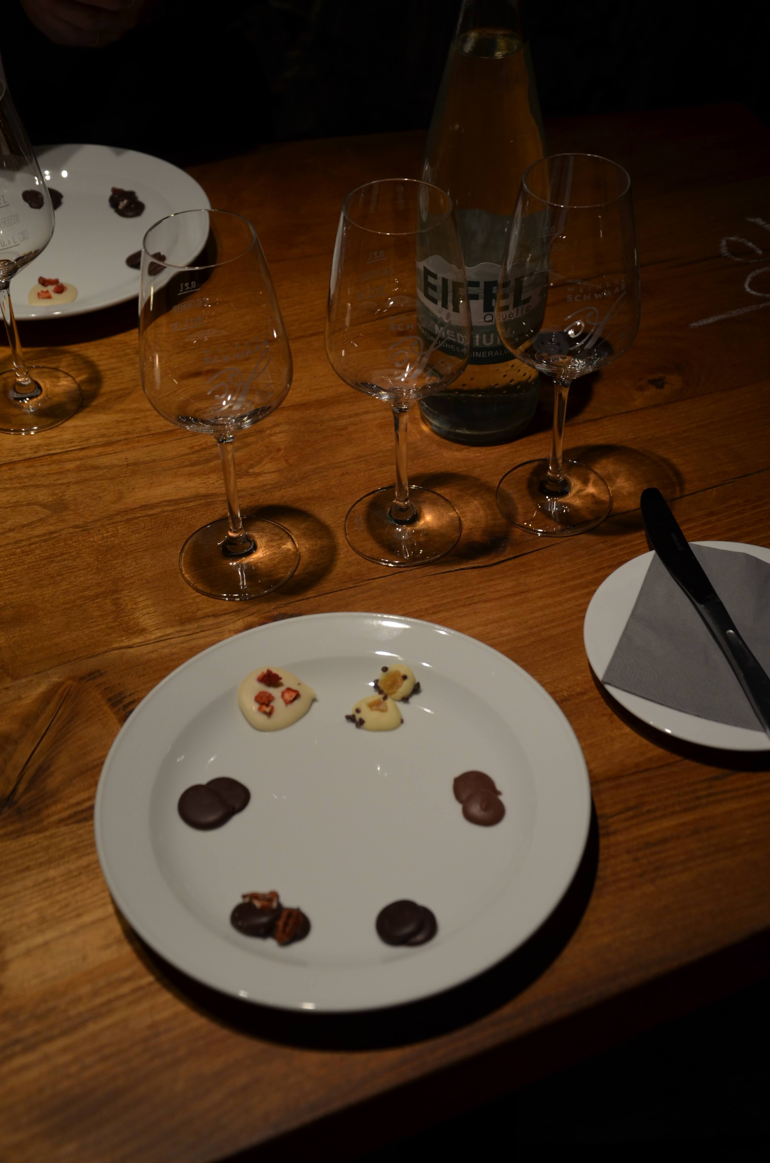 Blick auf drei Weingläser und Teller mit sechs verschiedenen Schokoladen während der Weinwerkstatt Wein und Schokolade im Weingut Schwaab in Koblenz. 